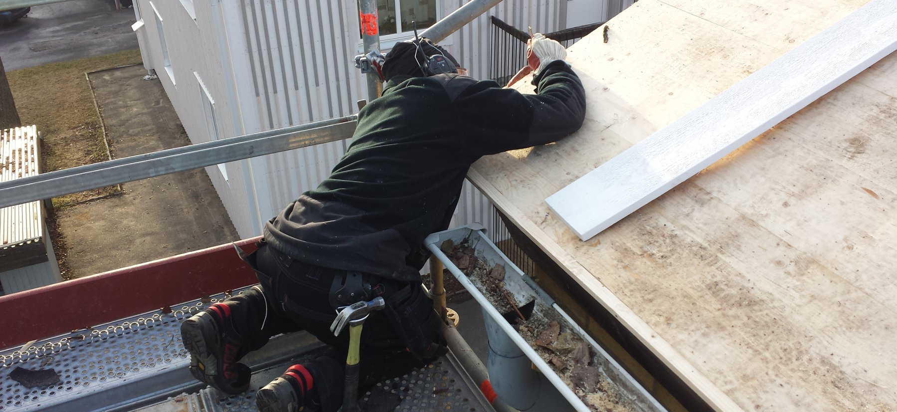 Bilden visar en snickare som sitter på en byggställning och mätar en takgavel under ett takbyte.